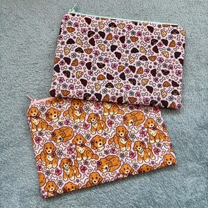 May include: Two rectangular zippered pouches. One features a pink background with a pattern of brown bones, flowers, and paw prints. The other has a pink background with a pattern of cartoon dogs, flowers, and bones. The zippers are light green and pink.