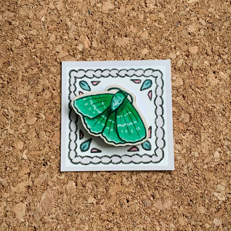 May include: A green wooden butterfly pin with a decorative border. The butterfly has a detailed design with a white outline.