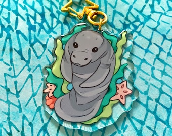 May include: A gray manatee cartoon character with a light blue background. The manatee is surrounded by green seaweed and pink and white seashells. The manatee is holding a blue and white seashell. The image is on a clear acrylic keychain with a gold star-shaped charm.