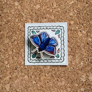 May include: A blue butterfly with brown accents is depicted on a white background with a decorative border. The butterfly is surrounded by green leaves and pink flowers.