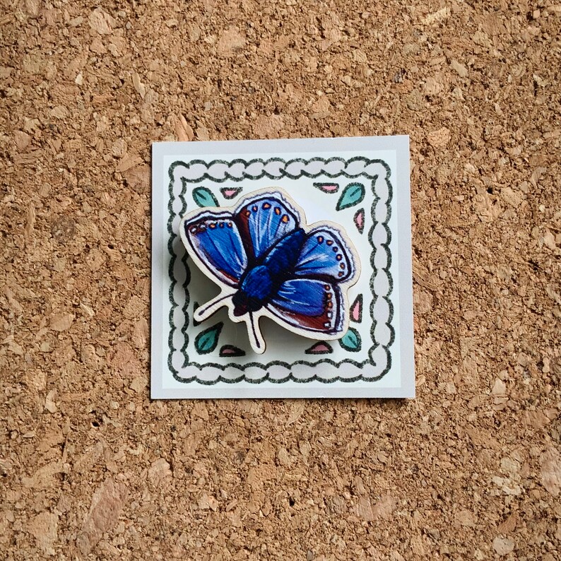 May include: A blue butterfly with brown accents is depicted on a white background with a decorative border. The butterfly is surrounded by green leaves and pink flowers.