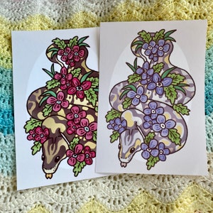 Peut inclure: Deux illustrations de serpents avec des motifs floraux. Le serpent de gauche a un motif jaune et brun avec des fleurs roses. Le serpent de droite a un motif jaune et violet clair avec des fleurs bleues.