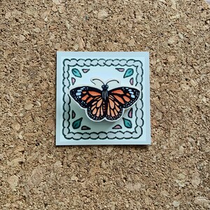 May include: A wooden butterfly pin with orange and black wings, surrounded by a decorative border with green leaves and pink flowers. The pin is on a white background.