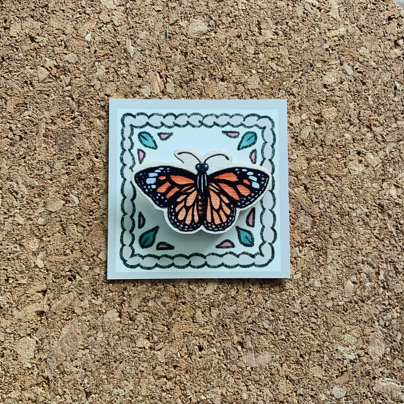 May include: A wooden butterfly pin with orange and black wings, surrounded by a decorative border with green leaves and pink flowers. The pin is on a white background.