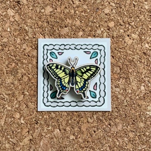 May include: A wooden butterfly pin with yellow, black, and blue wings, set against a white background with a decorative border. The butterfly is surrounded by small pink and blue flowers.