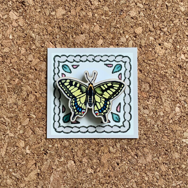 May include: A wooden butterfly pin with yellow, black, and blue wings, set against a white background with a decorative border. The butterfly is surrounded by small pink and blue flowers.
