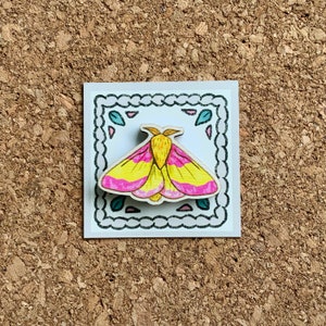 May include: A wooden pin featuring a hand-painted pink and yellow moth with black outlines. The pin is set against a white background with a decorative black border.