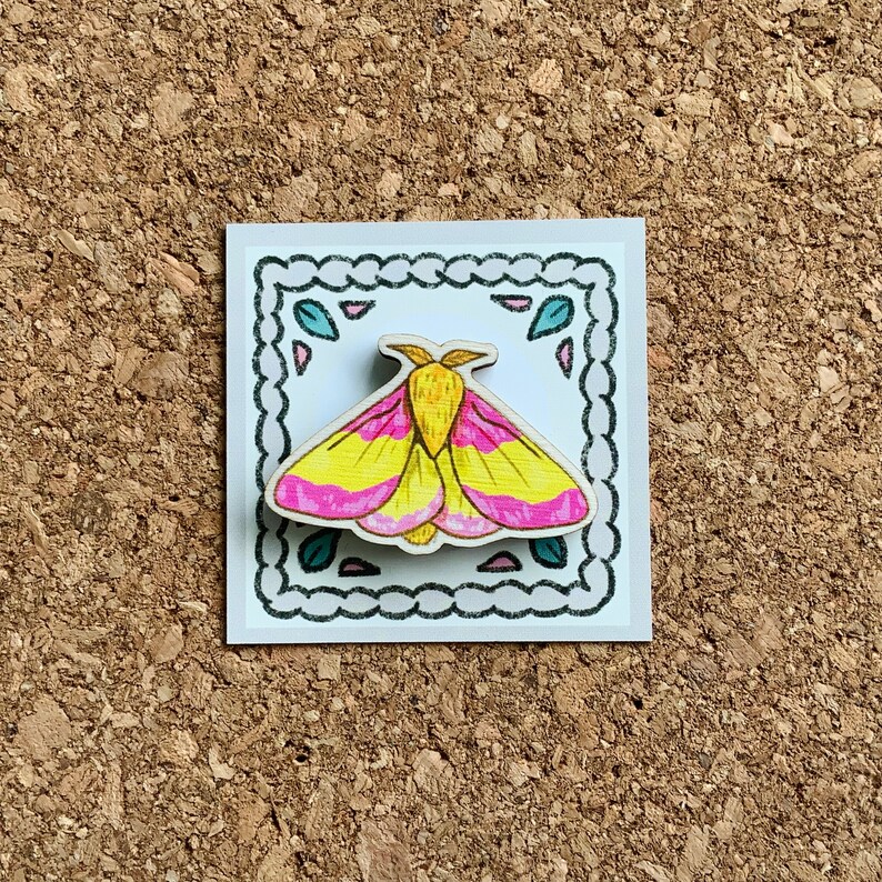 May include: A wooden pin featuring a hand-painted pink and yellow moth with black outlines. The pin is set against a white background with a decorative black border.