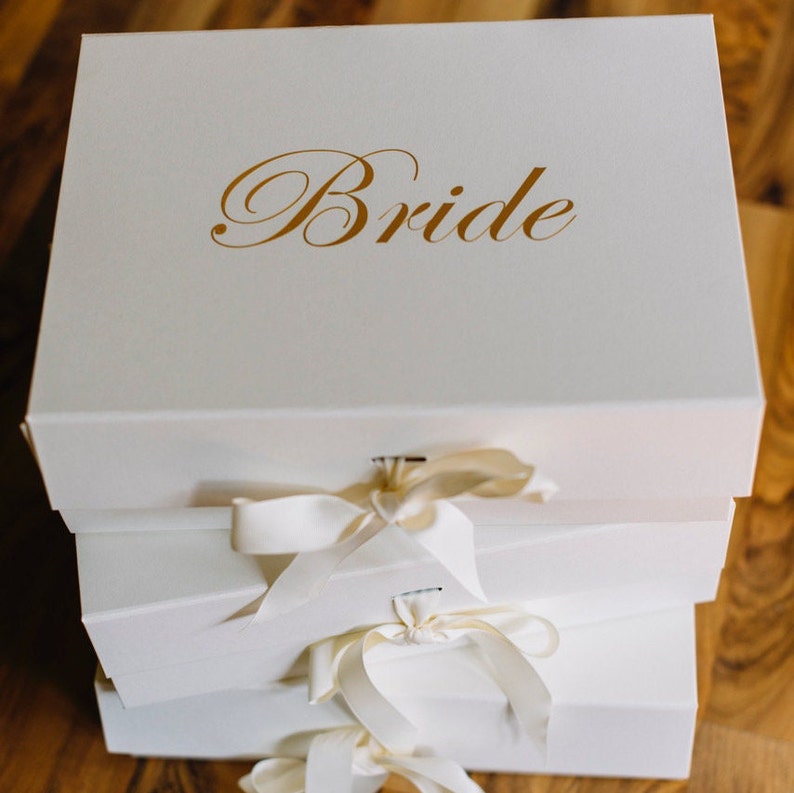 May include: A stack of three white gift boxes tied with white satin ribbon. The top box has the word "Bride" written in gold lettering.