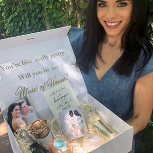 May include: A white gift box with a gold inscription that reads "You're like, really pretty. Will you be my Maid of Honor?" The box is filled with a variety of gifts, including a bottle of champagne, a mug, a rose, and a card.