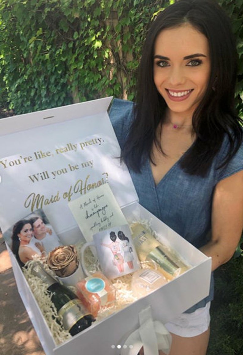 May include: A white gift box with a gold inscription that reads "You're like, really pretty. Will you be my Maid of Honor?" The box is filled with a variety of gifts, including a bottle of champagne, a mug, a rose, and a card.