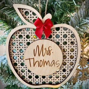Può includere: Un ornamento a forma di mela in legno con un motivo intrecciato e un fiocco rosso. L'ornamento ha un cerchio al centro con il testo "Mrs. Thomas" inciso su di esso.
