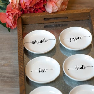 May include: Six white ceramic ring dishes with black cursive names: Amanda, Jessica, Lisa, Bride, Natalie, and Tiffany. The dishes are arranged on a metal tray with a wooden frame, with pink flowers in the background.