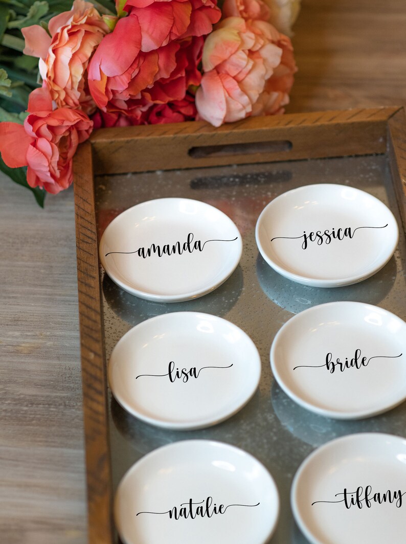 May include: Six white ceramic ring dishes with black cursive names: Amanda, Jessica, Lisa, Bride, Natalie, and Tiffany. The dishes are arranged on a metal tray with a wooden frame, with pink flowers in the background.