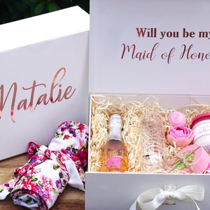 May include: White gift box with a pink and white floral robe, a bottle of champagne, a glass, a pink candle, and a pink soap. The box is decorated with the text "Will you be my Maid of Honor?" and the name "Natalie".