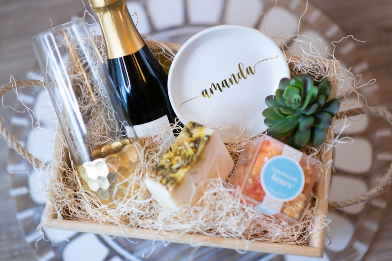 May include: A gift basket with a bottle of sparkling wine, a champagne flute, a white plate with the name "Amanda" in gold script, a bar of soap, a small succulent, and a box of champagne gummy bears. The basket is filled with straw.
