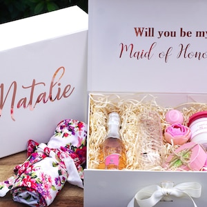 May include: White gift box with the words "Will you be my Maid of Honor" on the lid. The box is open and contains a floral robe, a bottle of champagne, a glass, a pink bath bomb, a pink flower, and a small jar of lotion.