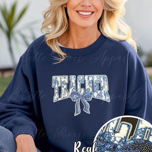 May include: Navy blue sweatshirt with the word "TEACHER" in a floral design and a sequined bow. The sweatshirt is a crew neck style. The words "Real Sequins" are printed below the bow.