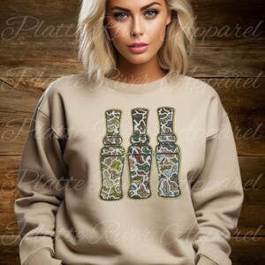 Puede incluir: Una sudadera beige con tres diseños decorativos de llamadas de caza. Las llamadas presentan patrones de camuflaje en tonos verdes, marrones y beige. Las palabras "Platte River Apparel" están impresas en la imagen.