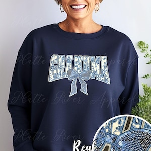 Puede incluir: Sudadera azul marino con la palabra "GRANDMA" en un diseño floral azul y blanco y un lazo de lentejuelas. Las palabras "Real Sequins" están escritas en blanco debajo.