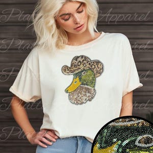Può includere: T-shirt color crema con un'anatra con paillettes che indossa un cappello da cowboy. L'anatra ha paillettes verdi, gialle e marroni. Il testo "Real Sequins!" è in basso nell'immagine. La maglietta è abbinata a jeans blu.