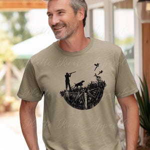 Puede incluir: Camiseta verde oliva con un gráfico negro de un cazador, un perro y pájaros. El diseño está dentro de una forma de brújula. La camiseta está hecha de un material suave y es adecuada para uso casual.