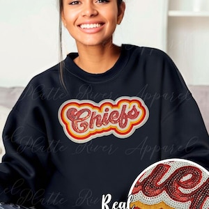 Puede incluir: Sudadera azul marino con el logo "Chiefs" con lentejuelas rojas, amarillas y blancas. El texto "Real Sequins" está en la parte inferior. La sudadera es de cuello redondo.