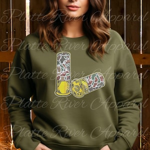 May include: Olive green sweatshirt featuring a graphic of two shotgun shells with a camouflage pattern. The shells have yellow and gold accents. The text "Platte River Apparel" is visible in the background.