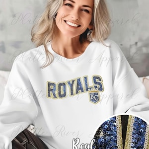 Puede incluir: Sudadera blanca con la palabra "ROYALS" en letras de lentejuelas azul marino y dorado. La sudadera también presenta un pequeño logotipo de KC. Las palabras "Real Sequins" están escritas en cursiva debajo.