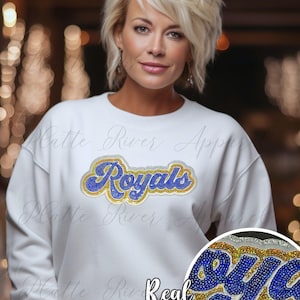 Puede incluir: Sudadera blanca con la palabra "Royals" en lentejuelas azules y doradas. La sudadera tiene cuello redondo y mangas largas. El texto "Real Sequins" está debajo.