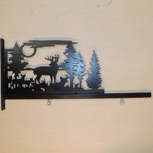 May include: Black metal wall-mounted sign featuring a silhouette of a deer, two trees, and a full moon. The sign has a horizontal bar with three rings for hanging.