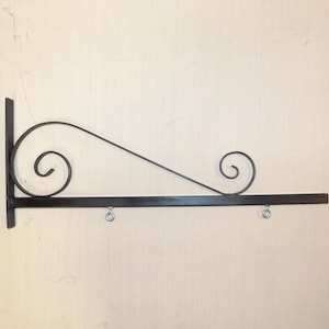 May include: Black metal sign holder with two decorative scrolls and three hanging rings. The holder is mounted on a white wall.