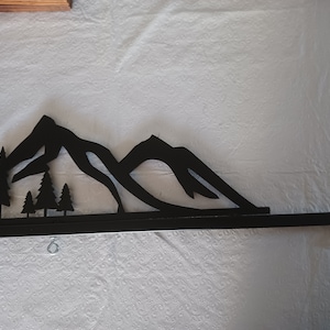 May include: Black metal sign bracket with a mountain and tree silhouette design. The bracket has two hanging rings.