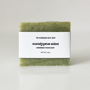 May include: A rectangular bar of handmade vegan soap, light green in color, with a white label wrapped around it. The label reads "THE BOHEMIAN BOX SHOP" above "eucalyptus mint" and "HANDMADE VEGAN SOAP". The net weight is 3.5 oz.