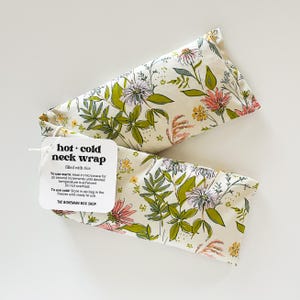 May include: Two cream-colored neck wraps with a floral pattern of pink, green, and yellow flowers and leaves. The wraps are labeled "hot + cold neck wrap" and filled with rice. A tag provides instructions for use.