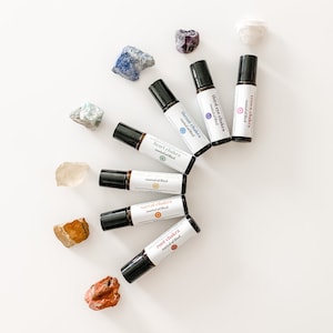 May include: An overhead shot features seven essential oil roller bottles labeled with chakra names, arranged in a semi-circle. Each bottle has a black cap and a white label with text and a colored chakra symbol. Various colorful crystals surround the bottles.