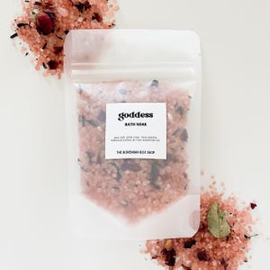 May include: A clear plastic pouch filled with pink bath salts, rose petals, and hibiscus petals. The pouch has a white label that reads "goddess BATH SOAK sea salt, pink clay, rose petals, hibiscus petals & rose essential oil THE BOHEMIAN BOX SHOP".