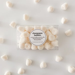 May include: A clear plastic box filled with white, round candies, scattered on a white surface. The candies are covered in white nonpareils. The box has a label that reads "champagne bubbles".