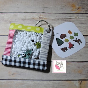 Puede incluir: Una bolsa de tela colorida con una ventana de plástico transparente llena de cuentas blancas. La bolsa tiene un borde a cuadros blanco y negro y una parte superior a cuadros rojo y blanco. Una tarjeta blanca con el texto "find me" e imágenes de criaturas del bosque está unida a la bolsa.