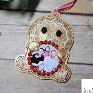 May include: A gold gingerbread man ornament with a red and white beaded frame holding a family photo. The ornament has a pink ribbon attached to the top.