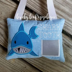 Könnte beinhalten: Ein blaues und graues Zahnfee-Kissen mit einem Hai-Design und dem Text "SHARK TOOTH". Das Kissen hat eine Tasche für einen verlorenen Zahn.