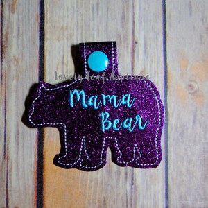 May include: A purple glittery bear-shaped applique with the words "Mama Bear" in turquoise script. The applique has a turquoise button and a small loop at the top. The background is a wooden surface.