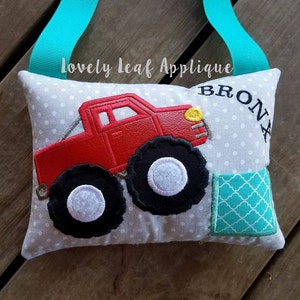 May include: A gray pillow with a red monster truck applique, a turquoise fabric square, and the name "BRONX" embroidered on it. The pillow has a turquoise ribbon attached.
