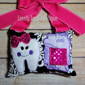 May include: A white tooth pillow with a pink bow, purple eyes, and a smiling mouth. The pillow is decorated with a black and white damask pattern and a purple and pink floral design with a pink sequin patch. The pillow has the name "Sydney Joy" embroidered on it. The pillow is tied with a pink ribbon with white stitching.