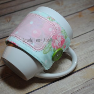 Pode incluir: Uma caneca de cerâmica branca com um aconchegante de caneca de tecido floral. O aconchegante tem um tecido de bolinhas rosa e branco com uma borda azul claro e um botão verde. O texto "Lovely Leaf Appetites" está bordado no aconchegante.