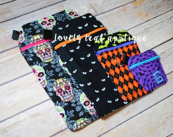 May include: Three different zippered pouches with Halloween-themed fabric. The largest pouch is black with a white eye pattern and features a skull and floral design. The middle pouch is black and orange with a checkered pattern. The smallest pouch is purple with a bat pattern and a blue letter G.