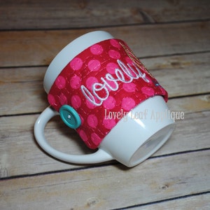 May include: White ceramic mug with a pink and white polka dot fabric mug cosy. The cosy has the word "lovely" embroidered on it in white thread. The mug cosy has a turquoise button.
