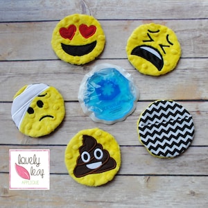 Può includere: Sei toppe rotonde in peluche in stile emoji con diverse espressioni, tra cui cuori, pianto, un bendaggio, cacca e uno schema a chevron. Le toppe sono su una superficie di legno.