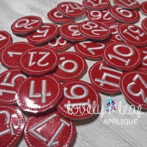 May include: Red felt circles with white numbers 1 to 24. The circles are decorated with a white stitched border. The circles are arranged in a random pattern on a wooden surface. The text "lovely leaf applique" is visible in the lower right corner of the image.