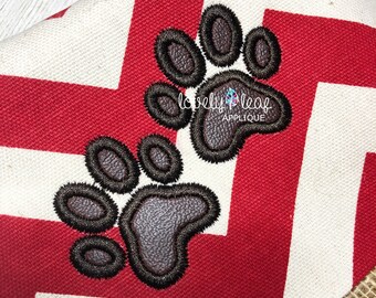 May include: Two brown paw prints with black outlines are embroidered on a red and white chevron fabric. The fabric is a close-up of a larger piece of fabric. The paw prints are a design element for a craft project.